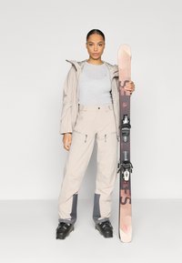 Suusapükste komplekt koos beežist veekindlast jope ja pükstega, halli särgi, mustade suusapaberi ning roosakas-pruunide suuskadega, millel mustad kinnitused.