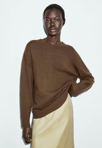 Donna con i capelli corti che indossa un maglione di lana marrone e una gonna beige, in piedi con una mano sul fianco contro uno sfondo chiaro e semplice.