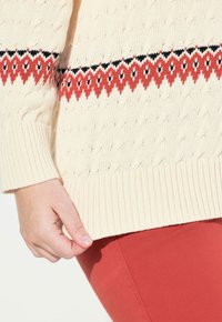 Maglione a maglia di colore crema con motivo a treccia e un design geometrico rosso e nero all'orlo. Polsini e bordo inferiore a coste.
