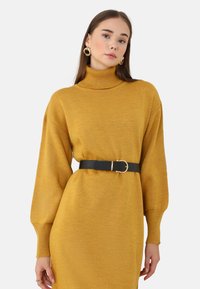 Vestito a maglia color giallo senape con collo alto a costine, maniche lunghe e una cintura nera con fibbia dorata. Tessuto morbido e lavorato a maglia.