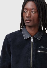 Jachetă din catifea în albastru închis, cu un guler din denim în contrast, fermoar la față și un mic buzunar pe piept cu detalii din argint. Material texturat.