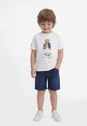 Νέο αγόρι που φοράει λευκό t-shirt με απεικόνιση ενός αρκουδάκι που κρατά ρακέτες τένις, τζιν σορτς και λευκά αθλητικά παπούτσια, στέκεται με τα χέρια στις τσέπες.