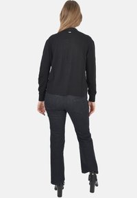 Cardigan nero in maglia con polsini a coste, abbinato a jeans a zampa d'elefante in denim scuro. L'outfit è completato da stivaletti nere con tacco.