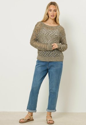 Vrouw staand, gekleed in een grofgebreide beige trui, opgerolde blauwe spijkerbroek en gouden sandalen met bandjes, tegen een effen lichte achtergrond.