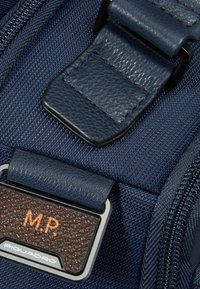 Marineblauw stof met een getextureerd donkerbruin leren patch. Voorzien van een metalen logo plaatje en een verstelbare leren riem met een gladde afwerking.