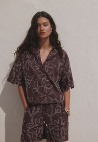 Femme aux longs cheveux foncés portant un haut à manches courtes sombre assorti et un short avec des motifs floraux abstraits blancs, debout contre un mur clair texturé.