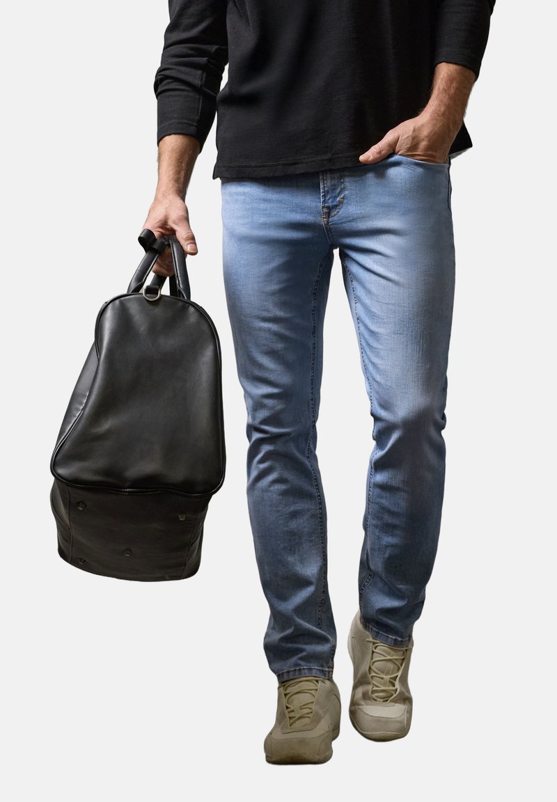 Un homme portant un jean bleu clair et des baskets beiges tient un sac à dos en cuir noir par les poignées, l'autre main dans la poche.
