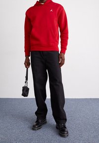 Felpa rossa con cappuccio e tasca frontale, con logo ricamato, abbinata a pantaloni neri larghi e scarpe nere. Tiene una piccola borsa nera con tracolla.