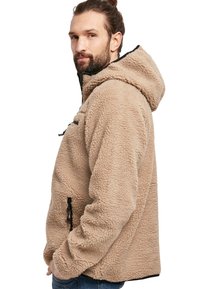 Chaqueta de forro polar beige con capucha, cremallera y detalles en negro. Presenta una superficie texturizada y bolsillos laterales con ajustes de cordón.