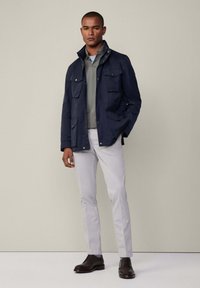 Chaqueta impermeable azul marino con un cuello alto, múltiples bolsillos frontales, llevada sobre una camiseta gris clara y combinada con pantalones gris claros y zapatos oscuros.