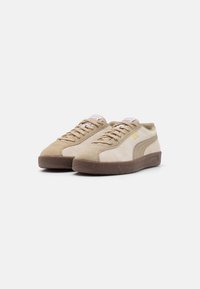 Baskets en suede beige avec une texture lisse, semelle en caoutchouc marron, design incurvé et lacets blancs, comportant des accents de marque discrets.