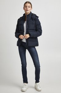 Giacca puffer blu navy con chiusura a zip, cappuccio removibile e due tasche anteriori. Abbinata a jeans scuri e sneakers bianche.