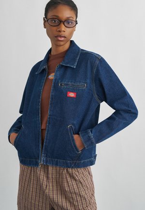 Tânără purtând ochelari ovali negri, jachetă denim albastru închis cu logo, cămașă maro și pantaloni în carouri, mâinile în buzunarele jachetei.