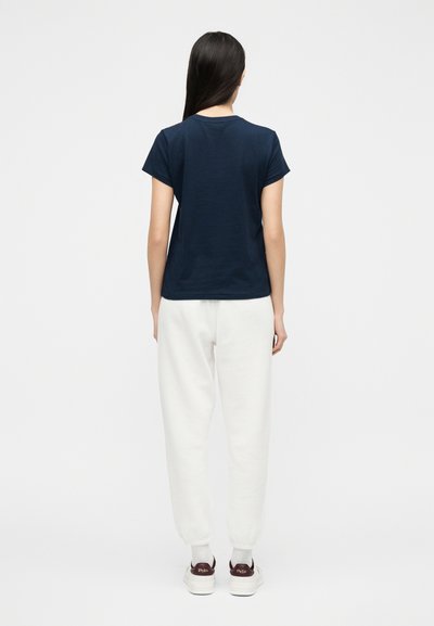 Γυναίκα με μακριά σκούρα μαλλιά, φορούσα μπλε σκούρο μπλουζάκι με κοντά μανίκια, λευκό παντελόνι jogger και λευκά αθλητικά, στεκόταν με την πλάτη προς τα εμπρός.