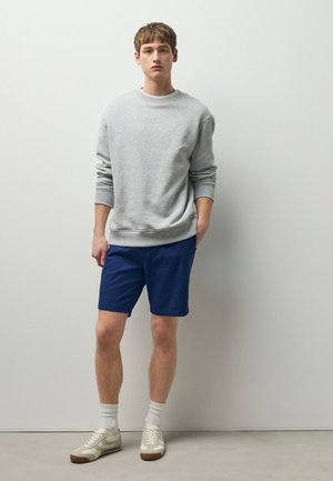 Junger Mann steht und trägt ein hellgraues Sweatshirt, blaue Shorts, weiße Socken und beige Sneakers vor einem einfarbigen hellen Hintergrund.