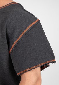 Donkergrijze t-shirt met ruwe snit aan de mouwen en onderkant. Oranje stiksels accentueren de halslijn en mouwen, met een zachte katoentextuur.