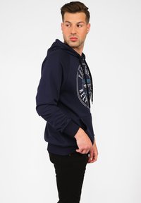 Marinblå hoodie med en framficka i känguru-stil, tryckt logotyp, dragsko-huvud och ribbade mudd, bärs med svarta byxor.