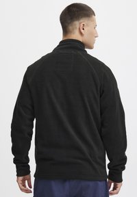 Schwarzer Fleece-Pullover mit hohem Kragen, Raglanärmeln und einer strukturierten Oberfläche. Verfügt über ein Diamantmuster am Hals und einen taillierten Saum.