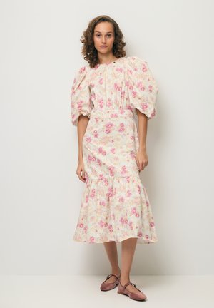 Kobieta stojąca przy ścianie, ubrana w białą sukienkę midi z różowym kwiatowym nadrukiem i bufkami, do tego różowe baleriny.