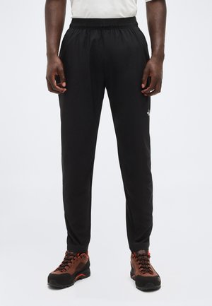 Muž v čiernych nohaviciach s elastickým pásom a v hnedých turistických topánkach stojaci s rukami uvoľnene pozdĺž tela na jednoduchom pozadí.