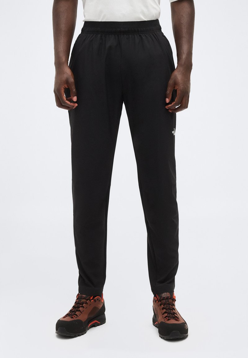 Άνδρας που φοράει μαύρο παντελόνι με ελαστική μέση και καφέ παπούτσια πεζοπορίας, στέκεται με τα χέρια χαλαρά στα πλευρά, σε απλό φόντο.