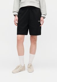 Άτομο που φοράει μαύρα σορτς Calvin Klein, λευκές κάλτσες, μπεζ αθλητικά παπούτσια και γκρι φούτερ με ριγέ μανσέτες.