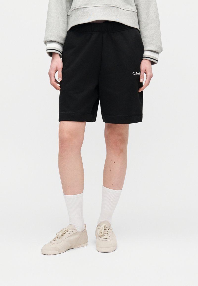 Άτομο που φοράει μαύρα σορτς Calvin Klein, λευκές κάλτσες, μπεζ αθλητικά παπούτσια και γκρι φούτερ με ριγέ μανσέτες.