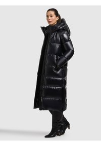 Lång svart dunjacka med blank finish, horisontell sömnad, överdimensionerad huva och sidofickor. Bärs med knähöga svarta stövlar.