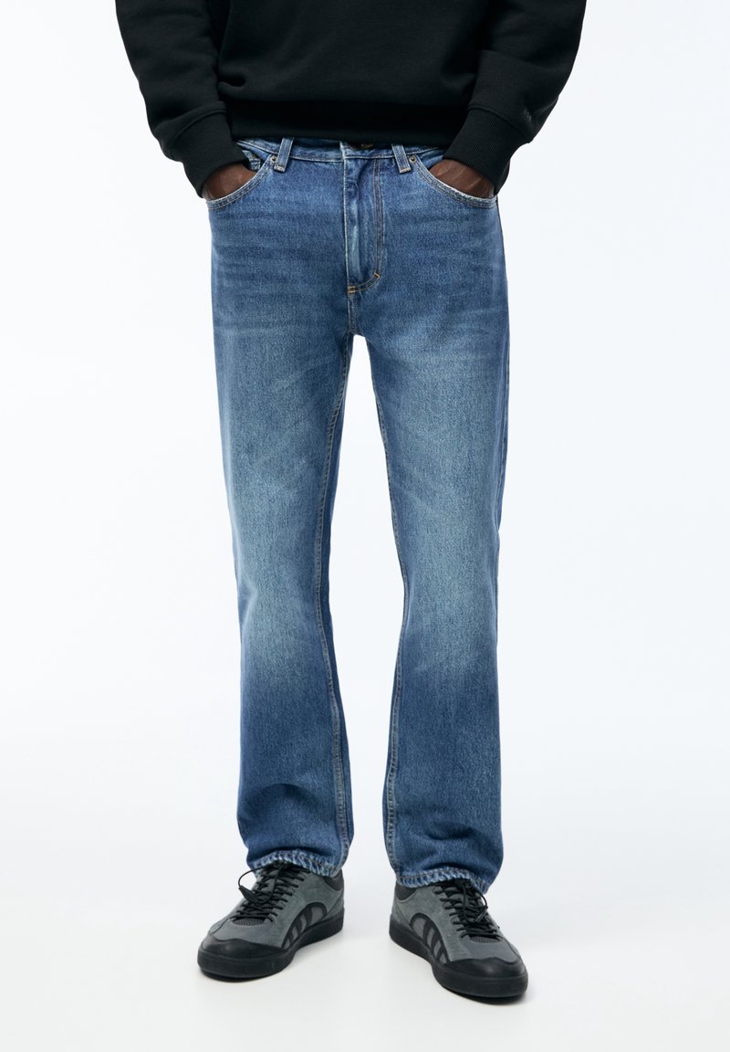 Personne portant un jean droit bleu, un sweat-shirt noir et des baskets gris-noir, debout les mains dans les poches.