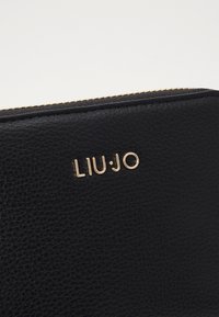 Svart teksturert lær lommebok med gullfarget "LIU•JO"-logo og glidelås langs toppen.