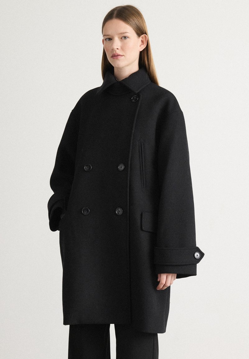 Cappotto di lana nera con colletto alto, design doppiopetto, grandi bottoni e tasche laterali. Presenta una vestibilità ampia e maniche con risvolto.