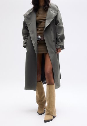 Personne portant un long trench-coat gris sur une courte robe marron, une large ceinture et de hautes bottes beiges en daim sur un fond blanc uni.