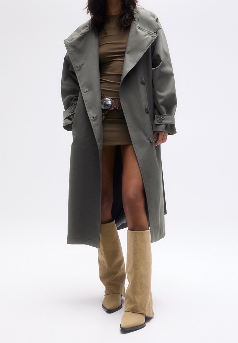 Personne portant un long trench-coat gris sur une courte robe marron, une large ceinture et de hautes bottes beiges en daim sur un fond blanc uni.