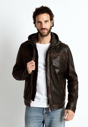 Homme aux cheveux bouclés et barbe portant une veste en cuir marron foncé avec capuche, un t-shirt blanc et un jean bleu sur fond blanc.