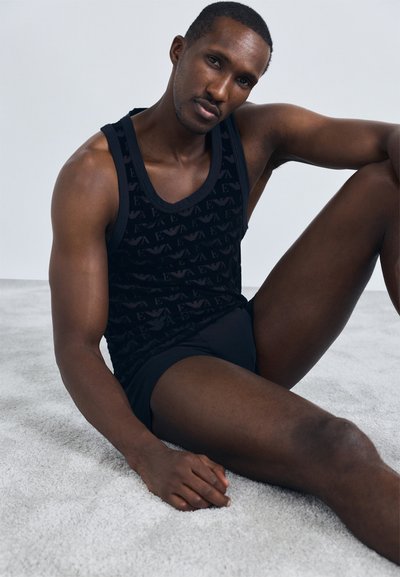 Sort ærmeløs tanktop med et struktureret mønster, parret med sorte shorts. Modellen sidder på et lysegråt tæppe og fremviser pasform og design.