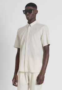 Camisa de lino de manga corta en color beige claro con botones en la parte delantera, combinada con pantalones a juego y gafas de sol negras de gran tamaño. Textura suave.