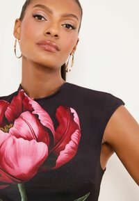 Čierny textilný top s veľkým ružovým kvetinovým vzorom, hladkou textúrou a priliehavým strihom s krátkymi rukávmi. Nosené zlaté kruhové náušnice.
