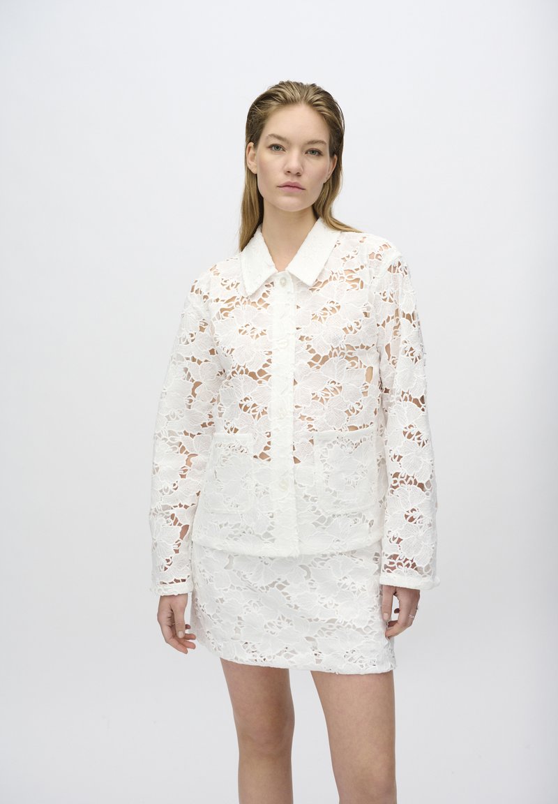 Conjunto de camisa y falda de encaje blanco que presenta patrones florales, un cuello con solapa y un cierre de botones, mostrando un diseño semitransparente.