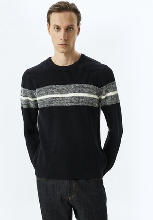 Jersey de punto azul marino con rayas horizontales grises y blancas en el pecho. Cuello redondo y diseño ajustado. Combinado con jeans oscuros.