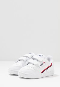 Vita sneakers med kardborreband, perforerade detaljer, röda och marinblå accenter, samt en distinkt logotyp på sidan. Gummisulor, klassisk design.