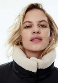 Zwarte pufferjas met een crèmekleurige, fluffy kraag, voorzien van een ritssluiting en zichtbare stikseldetails. Model heeft een neutrale expressie.