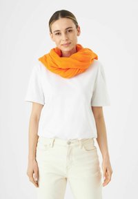 Écharpe orange et texturée enroulée autour du cou ; complétée par un t-shirt blanc uni et un jean de couleur claire.