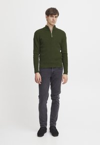 Maglione verde a zip con colletto alto, abbinato a jeans slim-fit grigio scuro e scarpe nere. Design minimalista, tessuto testurizzato.