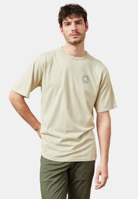 Camiseta de algodón beige con mangas cortas y cuello redondo, que presenta un logo circular verde en el lado izquierdo del pecho. Ajuste liso y relajado.
