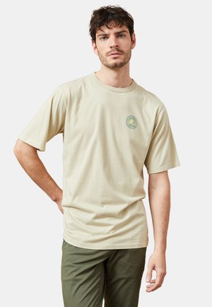 Camiseta de algodón beige con mangas cortas y cuello redondo, que presenta un logo circular verde en el lado izquierdo del pecho. Ajuste liso y relajado.
