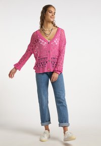 Mujer vistiendo un cárdigan de crochet rosa sobre una blusa blanca, jeans azul ajustados y remangados, zapatillas blancas y varias cadenas de cuentas de colores.