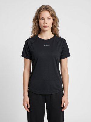 Ung kvinde med bølget blondt hår iført en simpel sort hummel sportst-shirt og sorte bukser, stående foran en hvid baggrund.