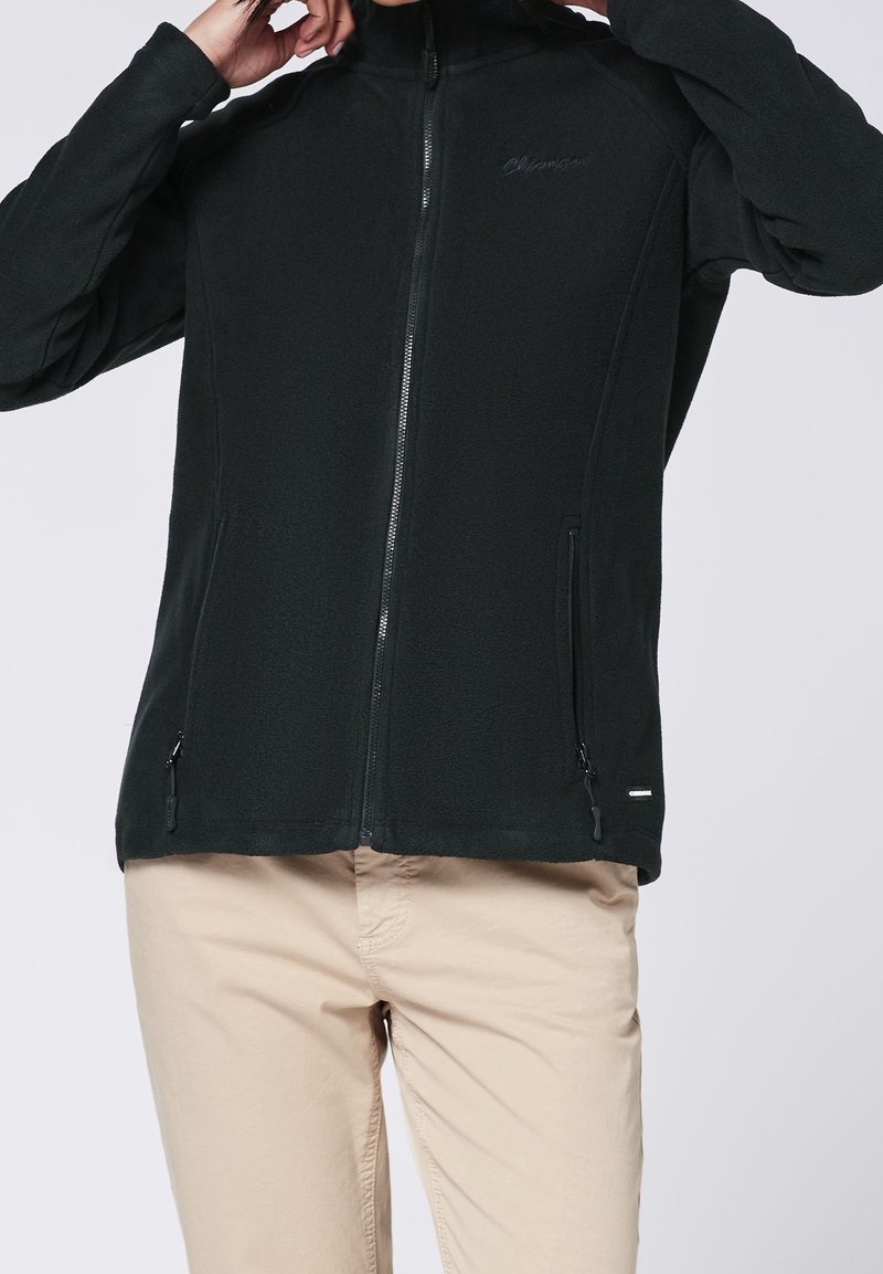 Personne portant une veste polaire noire zippée avec des poches zippées et un pantalon beige, ajustant le col de la veste.
