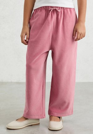 Gyermek rózsaszín, bőszárú, bordázott nadrágot visel, amelynek a derekán húzózsinór található, valamint bézs Mary Jane cipőt, és egy világosszürke padlón áll.