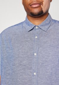 Košile s krátkým rukávem z modrého chambraye s límečkem na knoflíky, vybavená bílými knoflíky a jemně texturovanou tkaninou v uvolněném střihu.